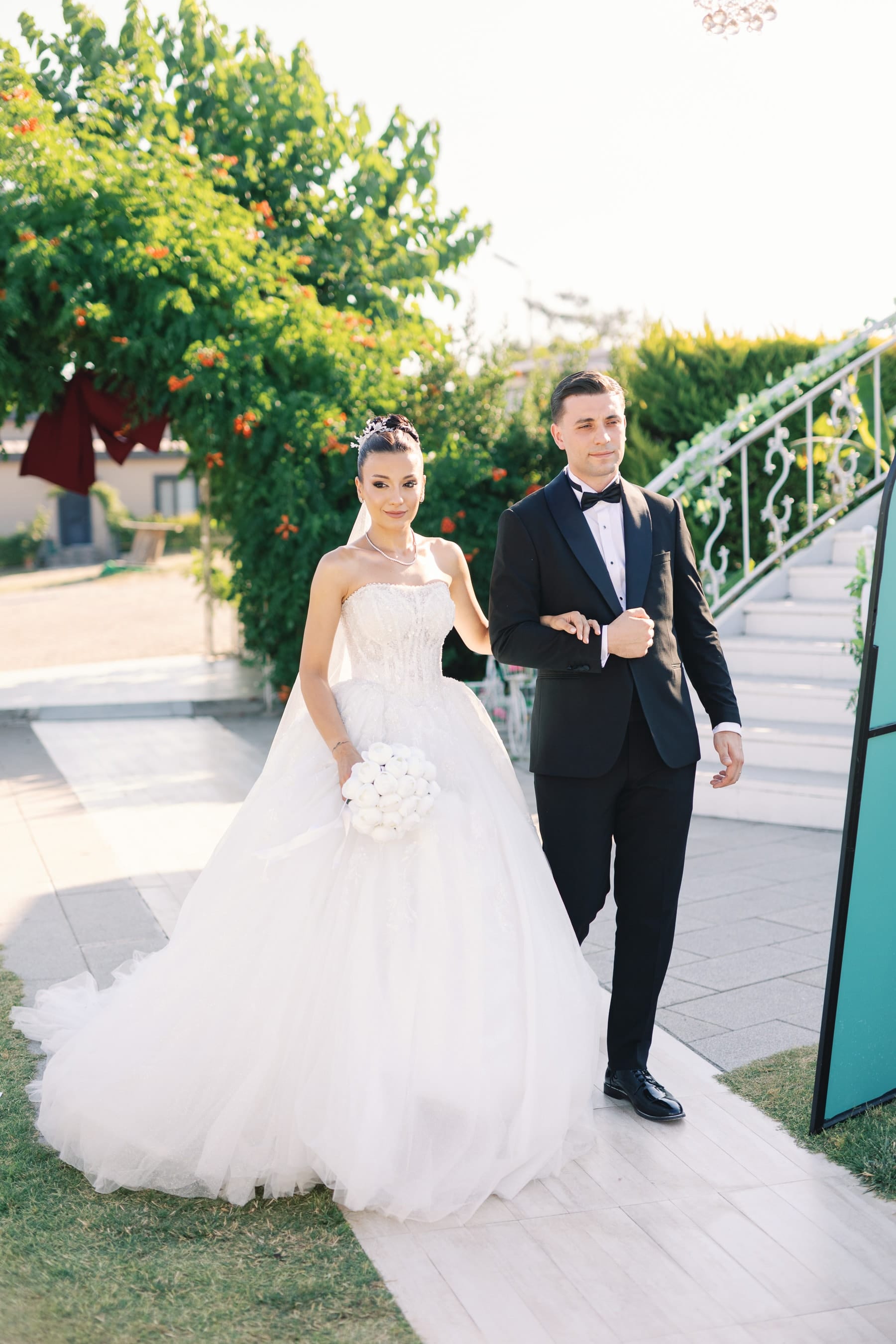 Bahçedeki beyaz merdivenin önünde kol kola yürüyen gelin ve damat