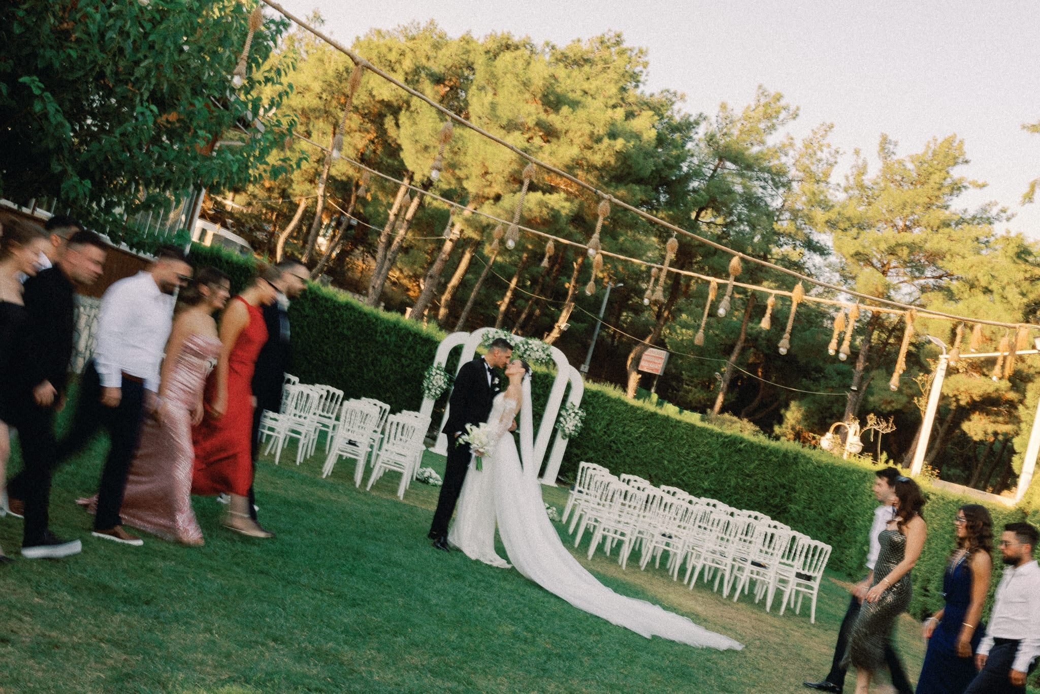 Çiçekli tören kemerinin önünde öpüşen gelin ve damat, iki yandan geçen davetliler